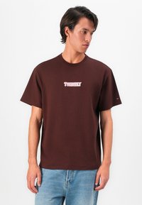Young man wearing a brown Tommy Hilfiger T-shirt and light blue jeans, looking sideways against a plain white background.