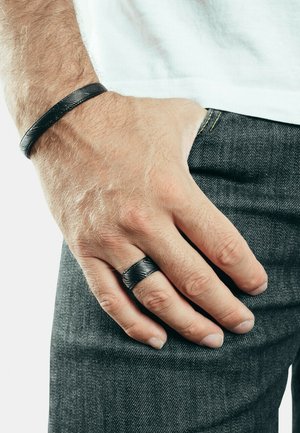 Black leather bracelet with textured surface on the wrist, paired with a matching black ring, worn with dark denim.