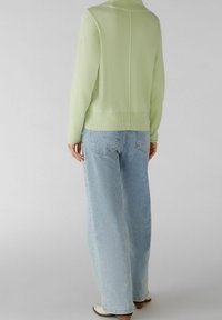 Persona che indossa un maglione verde chiaro e jeans blu chiaro a vita alta, in piedi davanti a uno sfondo grigio chiaro uniforme.