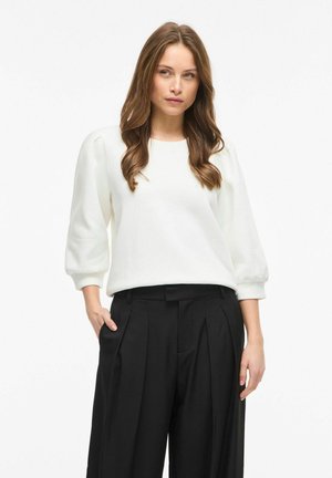 Femme aux longs cheveux bruns ondulés portant un chemisier blanc à manches bouffantes et un pantalon noir plissé taille haute, debout avec une main dans la poche.