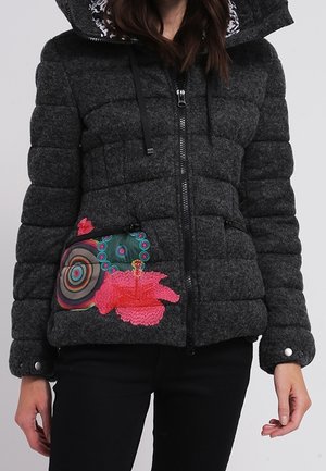 Mørkegrå quiltet jakke med hætte, frontlynlås og farverige broderede blomster- og cirkelmønstre på venstre side nær lommen.