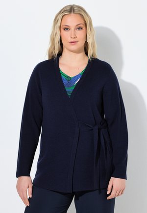 Kobieta w granatowym kardiganie zapinanym w pasie, nałożonym na niebiesko-zieloną wzorzystą bluzkę, stojąca na białym tle.