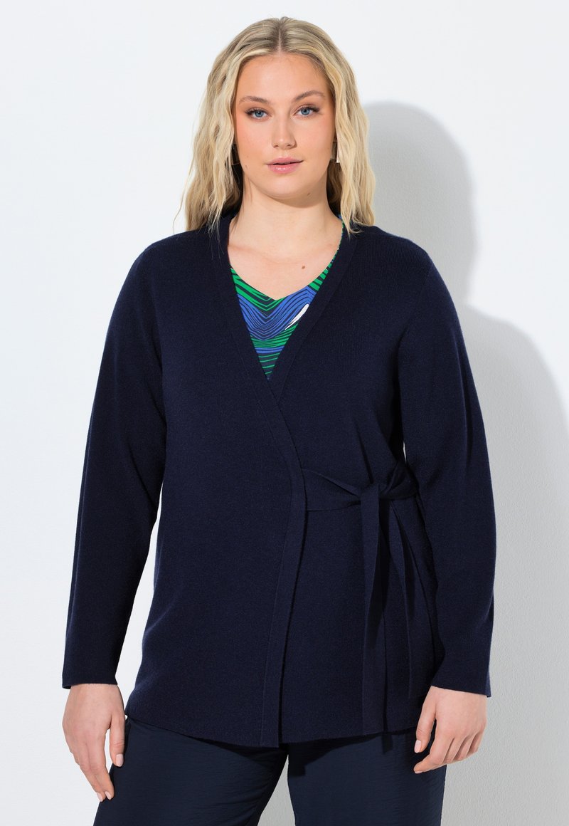 Frau trägt eine marineblaue Wickel-Strickjacke, die in der Taille gebunden ist, über einem blau-grün gemusterten Oberteil, stehend vor einem weißen Hintergrund.