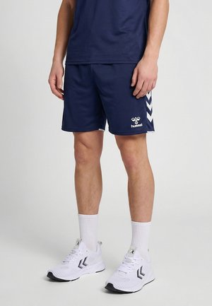 Person trägt marineblaue Sportshorts und Shirt, weiße Kniestrümpfe und weiße Sportschuhe und steht auf einem hellgrauen Boden.