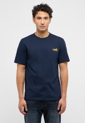 Homme aux cheveux bouclés portant un t-shirt Mustang bleu marine et un jean foncé, debout devant un fond gris clair uni.
