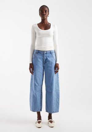 Kvinde står iført en hvid tætsiddende bluse med lange ærmer, løsthængende lyseblå denimjeans og hvide flade sko mod en ensfarvet hvid baggrund.