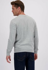 Man die met zijn rug naar de camera staat, gekleed in een lichtgrijze lange mouwen trui en donkerblauwe jeans met gele stiksels op de achterzakken.