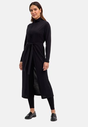 Donna che indossa un lungo cardigan nero con cintura, un dolcevita nero, leggings neri e mocassini neri, in piedi contro uno sfondo neutro.