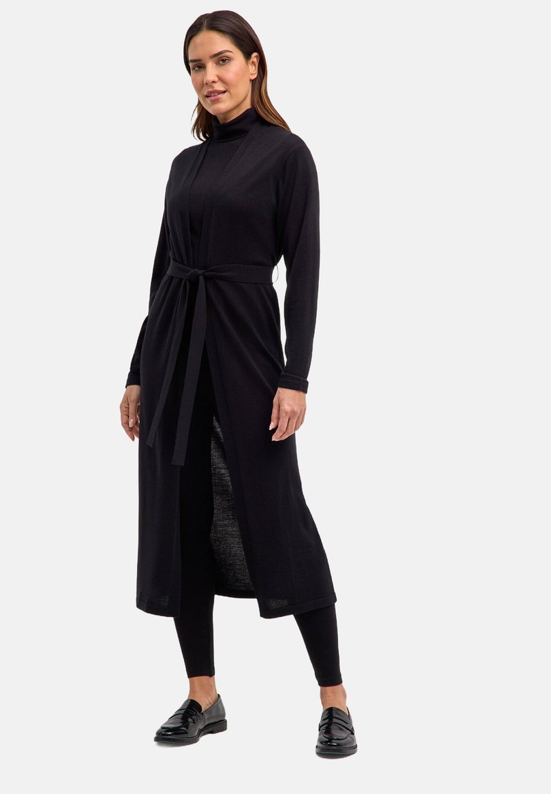 Kobieta w długim czarnym kardiganie z paskiem, czarnym golfie, czarnych legginsach i czarnych loafersach, stoi na jednolitym tle.
