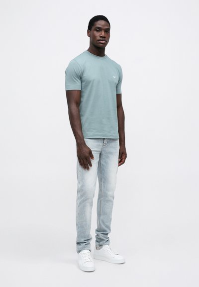 Mann står iført en grønn kortermet T-skjorte, lyseblå jeans og hvite joggesko mot en ensfarget hvit bakgrunn.