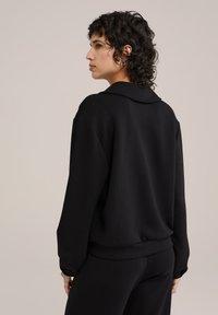 Fekete pulóver, enyhén túlnőtt fazonnal, ejtett vállakkal és hajtott gallérral. Sima textúra, illeszkedő nadrággal párosítva.