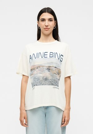 Femme aux longs cheveux foncés porte un t-shirt blanc oversize Anine Bing avec un paysage désertique, associé à un jean bleu clair.
