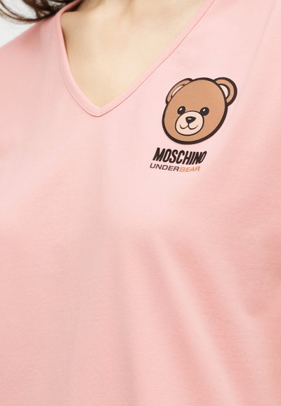 Ροζ ανοιχτό μπλουζάκι με V-λαιμόκοψη, με ένα μικρό καφέ γραφικό αρκουδάκι και το κείμενο «MOSCHINO UNDERBEAR» στο πάνω μέρος του στήθους.
