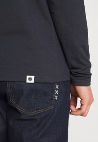 Mörkblå skjorta med lång ärm och en vit etikett vid fållen, kombinerad med mörka jeans i denim som har distinkt vit sömnad på bakfickan.