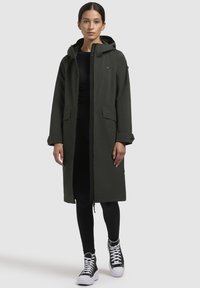 Lange donkergroene waterdichte jas met een capuchon, twee voorzakken en een ritssluiting. Draagt over zwarte leggings en sneakers.