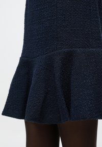 Obleka temno modre barve z rahlo razširjenim robom. Tkanina je videti kot tkalna z rahlo izbočeno površino, v kombinaciji s temnimi nogavicami.