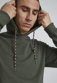 Olive green hoodie with a drawstring hood, featuring multicolored braided cords and a small logo patch on the front. Soft fabric texture.
