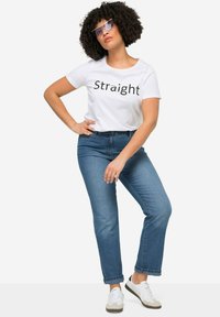 T-shirt bianco con la scritta "Straight" in nero, abbinata a jeans blu e sneakers bianche. Il modello ha i capelli ricci e indossa occhiali da sole.