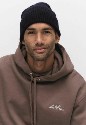 Brun hoodie med dragsnören, broderad text "Les Deux", och ribbad marinblå mössa. Hudtonen är varm, med ett subtilt skägg och kort hår.