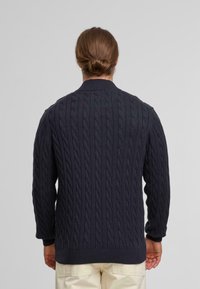 Donkerblauwe kabelgebreide trui met een hoge kraag en geribde manchetten, featuring een gestructureerd patroon, gemaakt van zacht materiaal, weergegeven vanuit de achterkant.