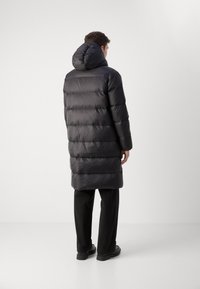 Schwarze Steppjacke mit Kapuze, mit einem gesteppten Design, langer Länge und elastischen Bündchen. Getragen über schwarzen Hosen und Schuhen.