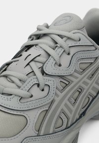 Gros plan d'une chaussure de sport gris clair avec des lacets croisés et des motifs de surcouche cousus sur un tissu en mesh respirant.