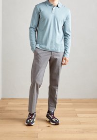 Světle modrá polokošile s dlouhými rukávy, šedé upravené kalhoty a tmavé tenisky s vícebarevnými akcenty. Pozadí z dřevěné podlahy.