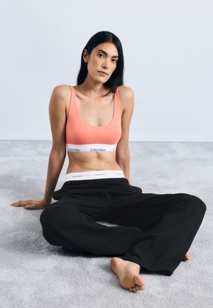 Top deportivo de color coral con tirantes anchos y banda blanca con el logo de Calvin Klein, combinado con pantalones negros holgados. Modelo sentado sobre una superficie gris claro.