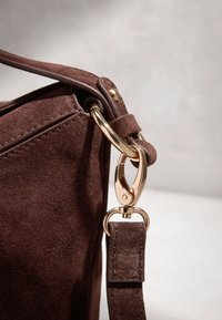 Gros plan sur un coin de sac en daim marron avec un fermoir métallique doré attachant une sangle en daim assortie sur un fond clair.