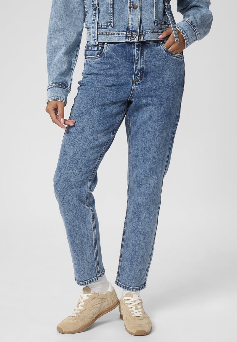 Jeans din denim albastru cu o croială dreaptă și textură estompată. Au cinci buzunare și cusuturi contrastante. Asortați cu adidași bej deschis.
