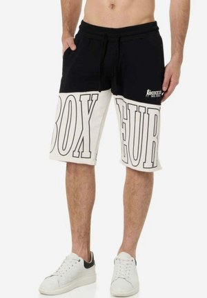 Mann trägt schwarz-weiße knielange Shorts mit großem "BOXEUR" Schriftzug und Logo, kombiniert mit weißen Sneakers.