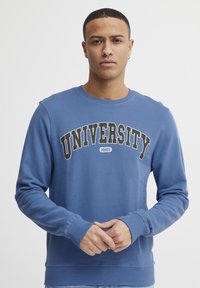 Sudadera azul con puños y cuello de canalé, con "UNIVERSITY" en letras negras en negrita cruzando el pecho. Material suave con un corte relajado.