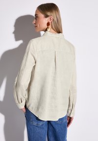 Camicia di lino beige chiaro con maniche lunghe, dettagli con piega sul retro e polsini con bottoni, abbinata a pantaloni in denim blu.