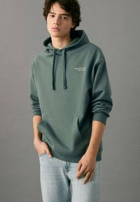 Sudadera con capucha en verde, tela suave con un bolsillo frontal, con el texto "CHILLIN' IN THE LOBBY" en blanco. Combinada con jeans claros.