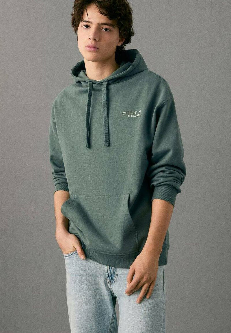 Sudadera con capucha en verde, tela suave con un bolsillo frontal, con el texto "CHILLIN' IN THE LOBBY" en blanco. Combinada con jeans claros.