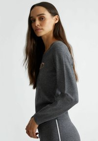 Maglione grigio con scollo rotondo, maniche lunghe e un design decorativo a forma di cuore realizzato con piccole perle sul petto. Tessuto morbido.