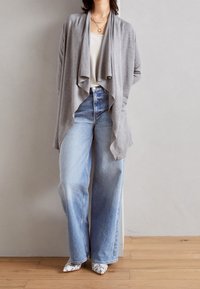 Gray cardigan with draped front, worn over a white top. Paired with light wash, wide-leg jeans and patterned pointed-toe shoes. Wood floor background.