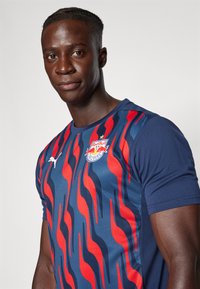 Kék navy sportpóló piros és kék hullámmintákkal, logóval és Puma emblémával. Rövid ujjak és érdes szövet.