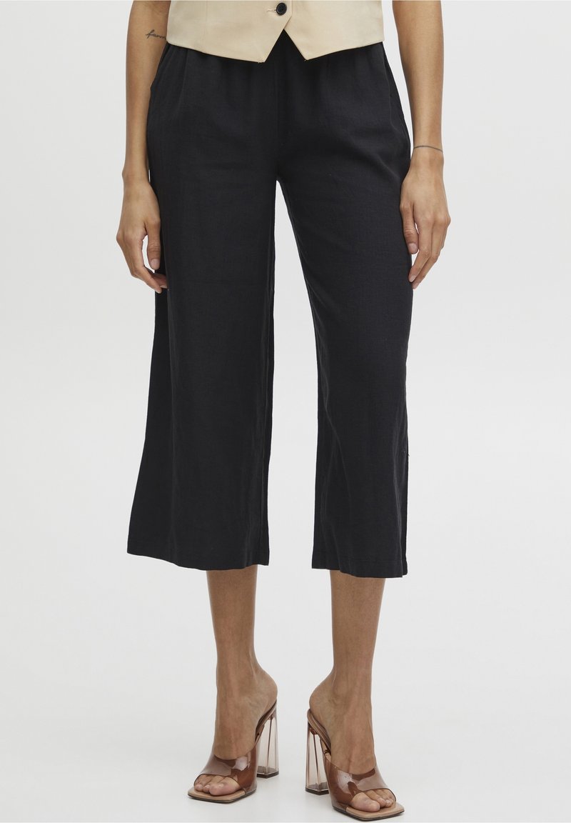 Personne portant un pantalon noir large et court, un haut beige sans manches et des sandales transparentes à talons hauts, debout devant un fond uni.