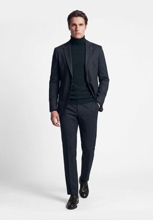 Homme portant un pull à col roulé noir, un costume sombre sur mesure et des chaussures habillées noires marchant avec une main dans la poche sur un fond uni.