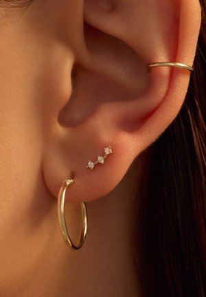 Goldene Creolen mit kleinen Cluster-Stiften, die mit drei runden klaren Steinen verziert sind. Glatte Metalloberfläche, glänzende Oberfläche und minimalistisches Design.