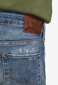 Bakfickan på blå denimjeans med blekta detaljer och en brun läderetikett från Jack & Jones, i kombination med en grön skjorta.
