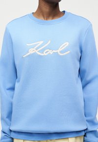 Άτομο που φοράει ανοιχτό μπλε φούτερ με λευκή εγχάρακτη επιγραφή "Karl" στο στήθος, συνδυασμένο με μπεζ παντελόνι.