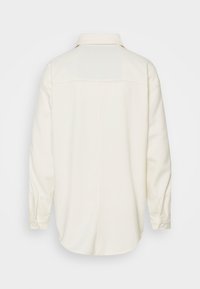 Vaalean beige pitkähihaisissa paidassa on kaulus, napitettu hihansuu ja kaareva helma, esitetty takaa yksinkertaisella valkoisella taustalla.