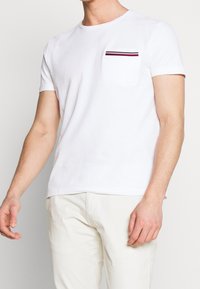 Biała bawełniana koszulka z okrągłym dekoltem i krótkimi rękawami, z lewą kieszenią na piersi ozdobioną poziomym detalem w czerwone, granatowe i białe paski.