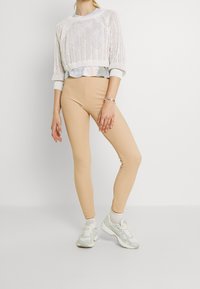 Beige legginsit, jotka on valmistettu joustavasta kankaasta, yhdistetty valkoiseen neulottuun lyhythihaiseen villapaitaan. Jalkineena kevyet harmaat lenkkarit.