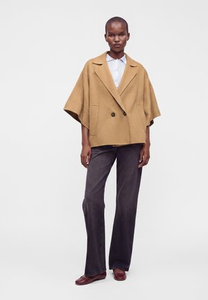 Femme portant un manteau court couleur camel sur une chemise blanche à boutons, un jean large foncé et des chaussures bordeaux, debout devant un fond uni.