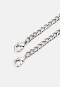 Stříbrný kovový řetěz s oválnými články a dvěma karabinkovými sponami. Povrch je lesklý a hladký, což ukazuje na leštěný vzhled.