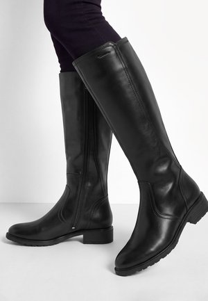 Bottes noires en cuir jusqu'aux genoux avec fermeture éclair latérale portées avec un pantalon ajusté foncé sur fond blanc.