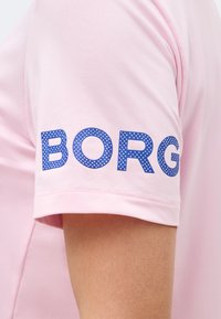 Ροζ πουκάμισο με κοντά μανίκια και μπλε πουα κείμενο "BORG" στο άνω μέρος του βραχίονα, φορεμένο από άτομο με ανοιχτόχρωμο τόνο δέρματος.